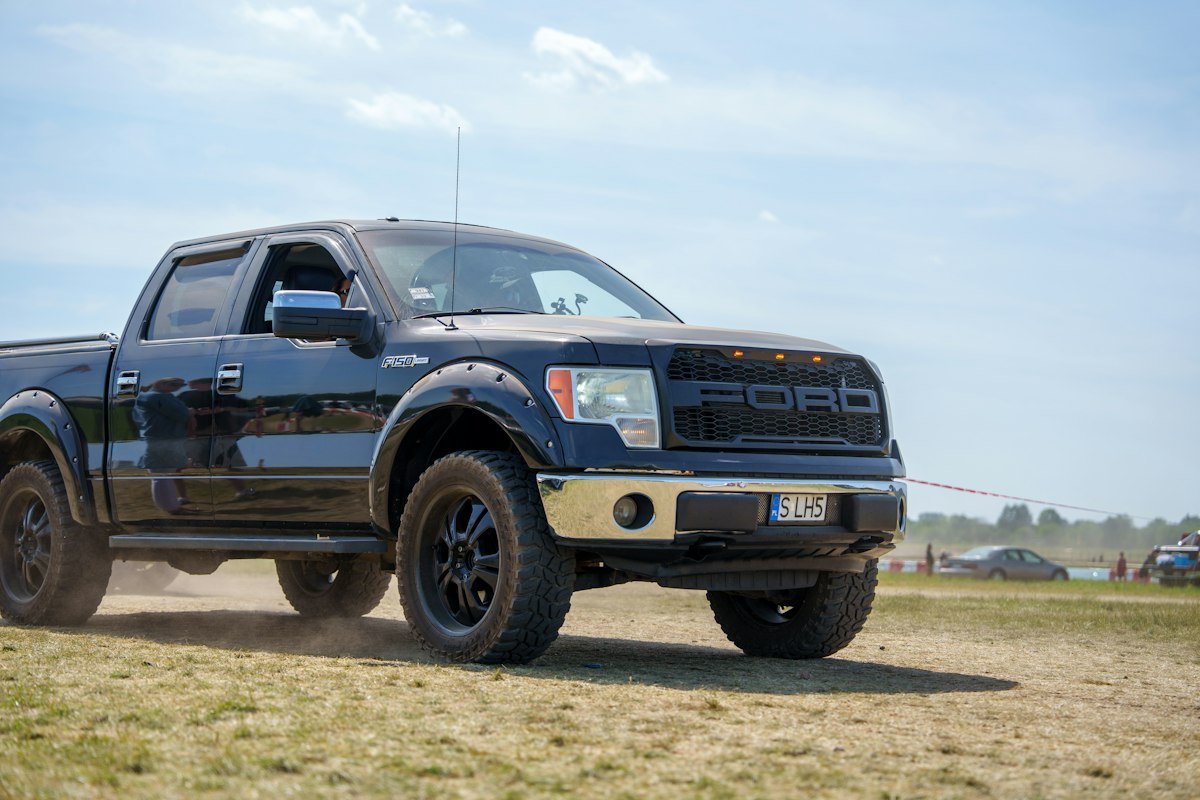 A dark Ford F-150 pickup truck kicking up dust as it drives across an unpaved field, representing the SUVs and trucks Ghanaian buyers import from US auctions